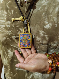 Tibetan thangka pendant with colorful deity painting, worn on black cord necklace, shown with Buddhist prayer beads on hand